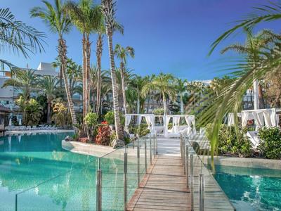 Holzsteg führt über einen Pool zu einer Lounge mit weißen Zelten und Palmen bei klarem Himmel.