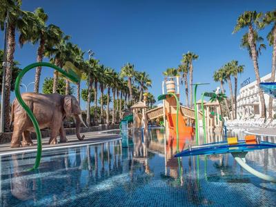 Děti si hrají v tropickém bazénu s vodními skluzavkami a palmami pod jasnou oblohou.