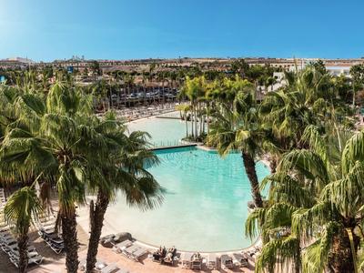 Velký bazén s palmami a lehátky v rekreačním areálu s výhledem na město v pozadí.