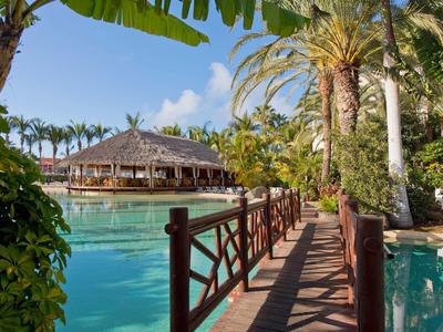 Dřevěná chodba s zábradlím vede k doškovému altánu nad tyrkysovou vodou obklopenou palmami.