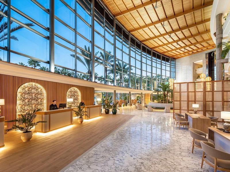 Prostorná hotelová lobby s vysokými skleněnými okny, rostlinami a moderní recepcí.