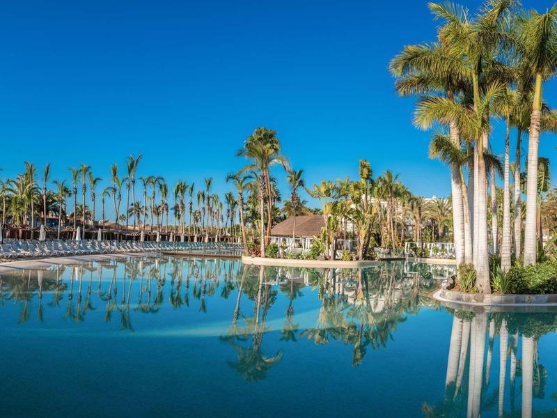 Velký hotelový bazén s palmami a jasně modrou oblohou v tropickém prostředí.