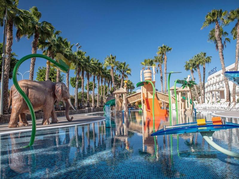 Děti si hrají v tropickém bazénu s vodními skluzavkami a palmami pod jasnou oblohou.