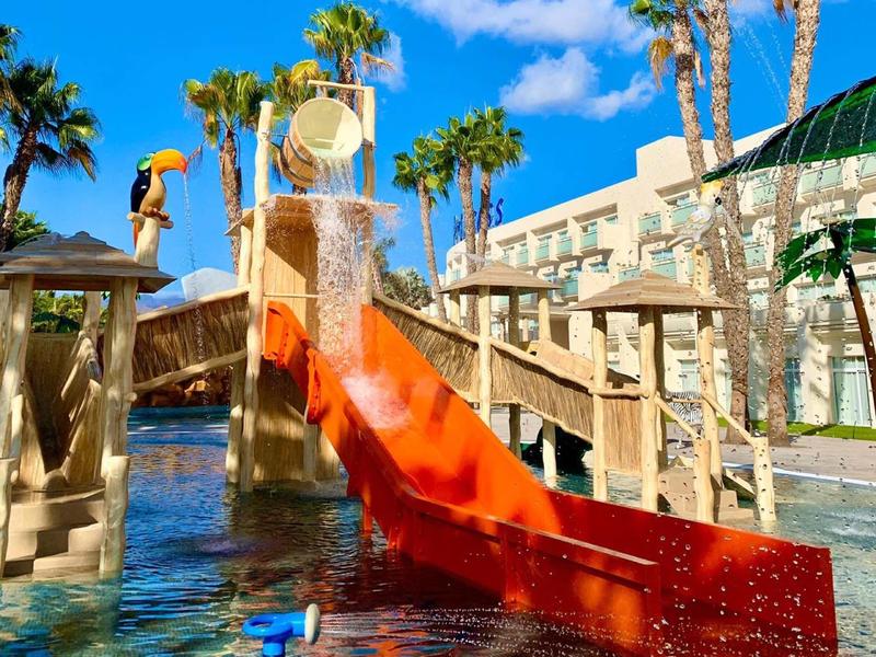 Vodní skluzavka s vodopádem v bazénové oblasti obklopené palmami a hotelovou budovou.
