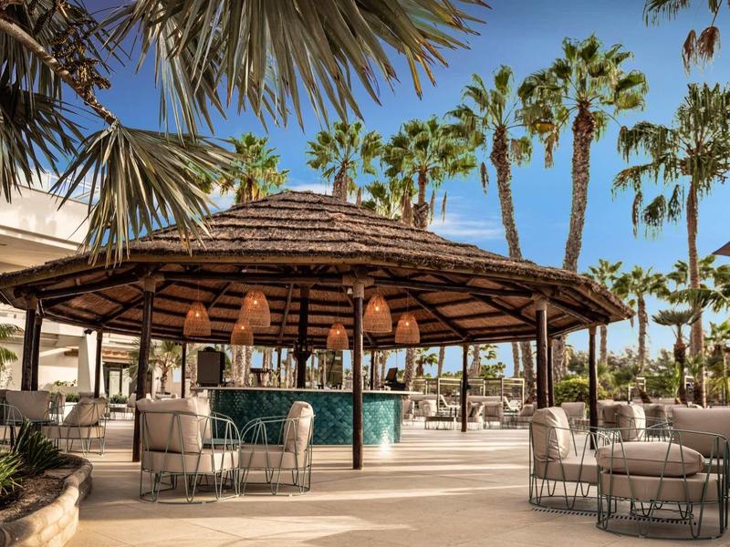 Otevřený bar se slaměnou střechou a posezením, obklopený palmami pod jasnou oblohou.