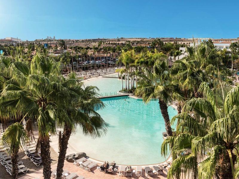 Velký bazén s palmami a lehátky v rekreačním areálu s výhledem na město v pozadí.