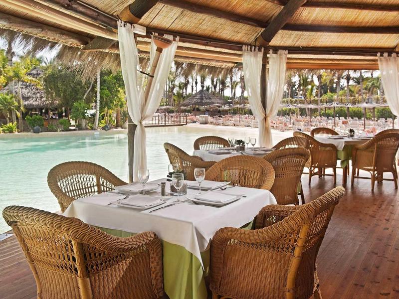 Zastřešená jídelní část s proutěnými židlemi vedle bazénu v tropickém letovisku.