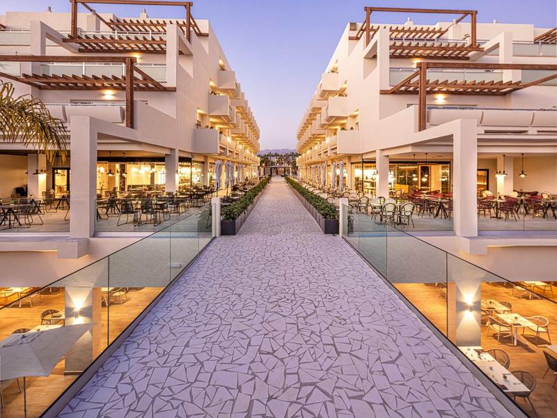 Moderní hotelový komplex se symetrickými budovami a venkovními restauracemi při západu slunce.