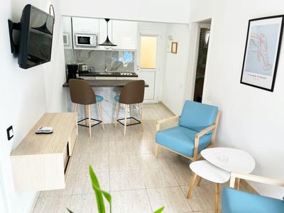 Helles Hotelzimmer mit moderner Küche, zwei Stühlen, blauem Sessel und Wandfernseher.