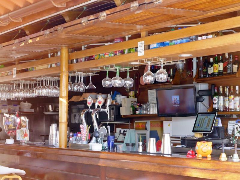 Espace bar avec comptoir en bois, tabourets, bouteilles de vin et spiritueux, et verres suspendus.
