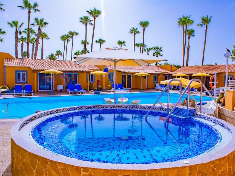 Bain à remous rond devant une grande piscine et des bungalows jaunes avec des palmiers.