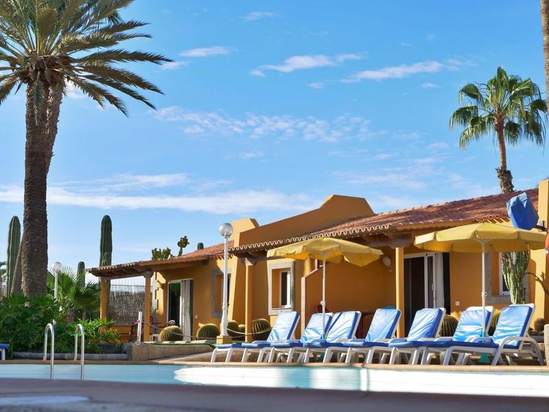 Piscine d'hôtel avec transats bleus, parasols et palmiers sous un ciel bleu.