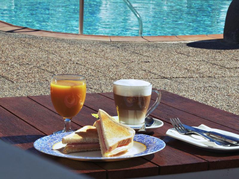 Table en bois avec petit-déjeuner composé de sandwich, café et jus près de la piscine.