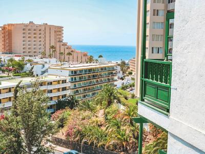 Pohled na hotelové budovy, zelené rostliny a modré moře v pozadí.