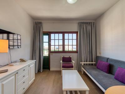 Světle zařízený hotelový pokoj s pohovkou, televizí, stolem a balkonem.