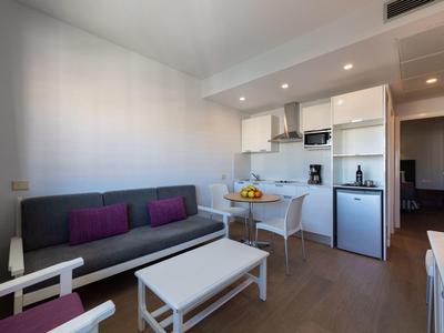 Moderní obývací prostor se šedou pohovkou, bílým stolem a malou kuchyňskou linkou v pozadí.