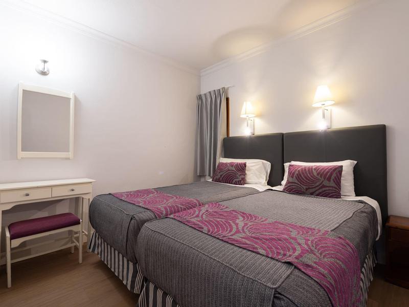 Moderně zařízený hotelový pokoj s manželskou postelí, psacím stolem a židlí v neutrálních barvách.