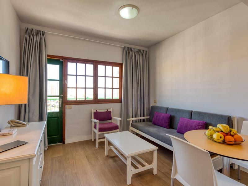 Příjemný hotelový pokoj s pohovkou, stolem, židlemi, ovocem a přístupem na balkon.