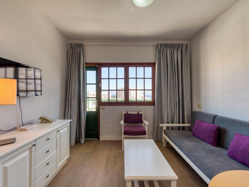 Světle zařízený hotelový pokoj s pohovkou, televizí, stolem a balkonem.