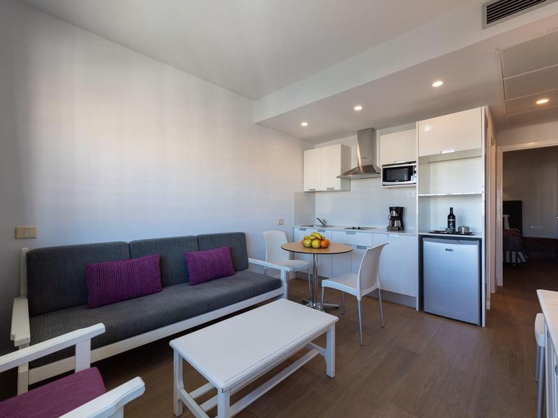 Moderní obývací prostor se šedou pohovkou, bílým stolem a malou kuchyňskou linkou v pozadí.