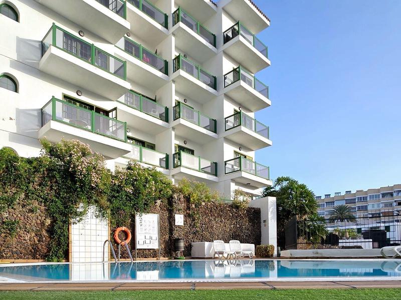 Moderní hotelový komplex s bazénem, lehátky a upraveným trávníkem pod jasně modrou oblohou