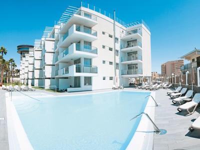 Moderner Hotelkomplex mit großem Pool, weißen Liegestühlen und Palmen bei klarem Himmel.