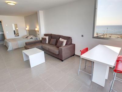 Lichte moderne hotelkamer met bank, bed, eettafel en zeezicht door groot raam.