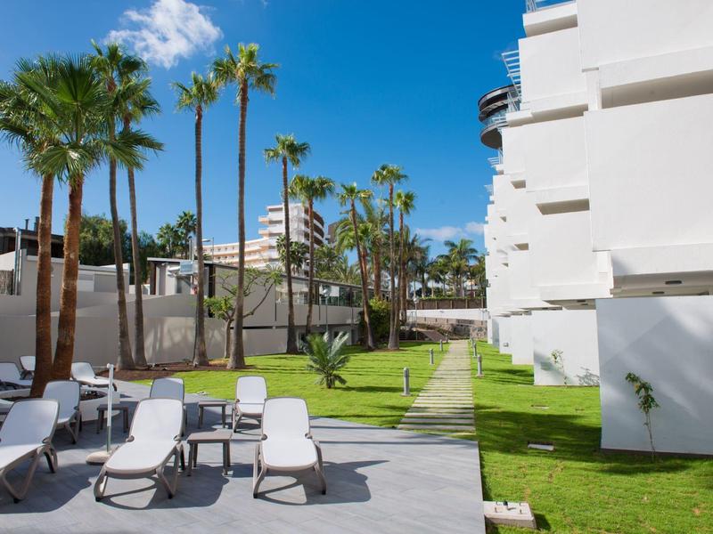 Moderne buitenruimte van een hotel met ligstoelen, palmbomen en goed onderhouden gras onder een blauwe lucht.