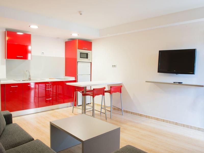 Moderne woonruimte met rode kitchenette, eettafel met stoelen en wandtelevisie.