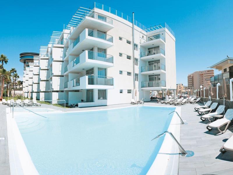 Modern hotelcomplex met groot zwembad en ligstoelen onder een heldere blauwe lucht.