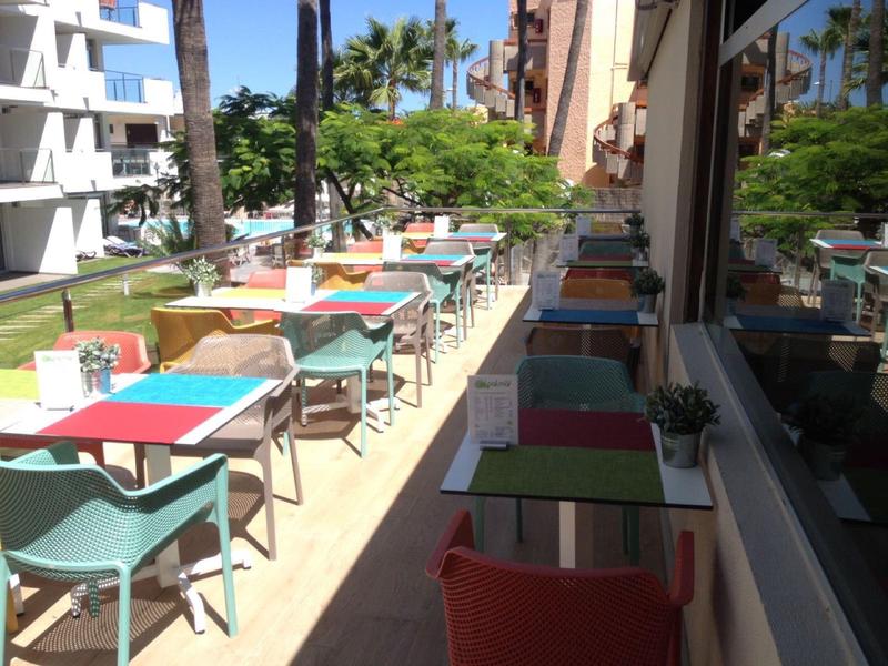 Zonnig terras met kleurrijke tafels en stoelen naast een hotelgebouw.