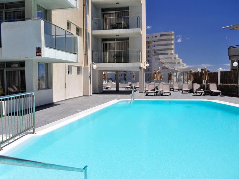 Modern buitenzwembad met ligstoelen naast een wit hotelgebouw onder een blauwe lucht.