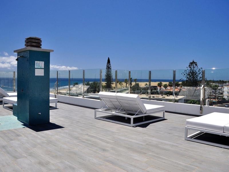 Dakterras met ligstoelen, glazen hek en uitzicht op omliggende gebouwen bij heldere lucht.