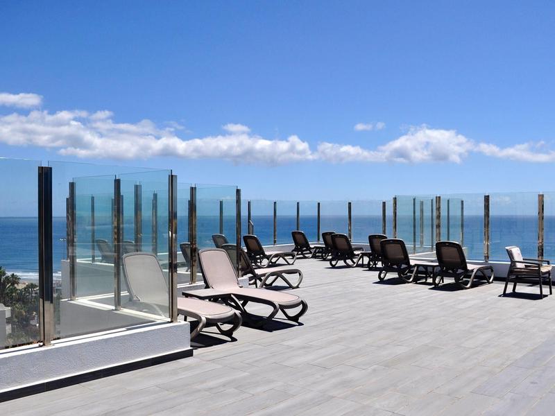 Dakterras met ligstoelen en glazen balustrade met uitzicht op zee onder een blauwe lucht.