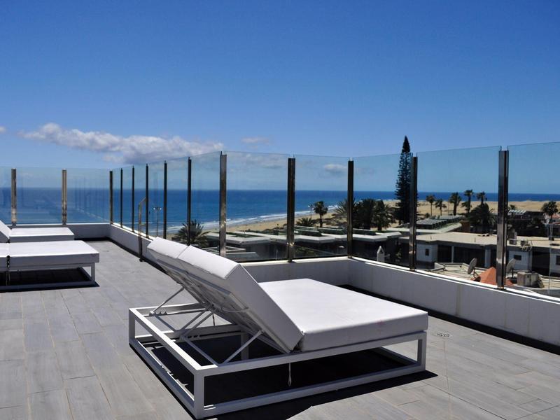 Moderne ligstoelen op een dakterras met glazen balustrade en uitzicht op zee onder een blauwe lucht.