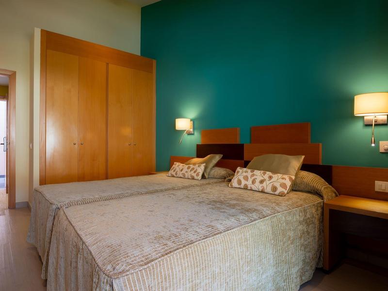 Hotel room with twin beds, wooden wardrobe, teal accent wall, and wall-mounted lamps.