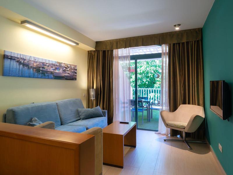 Cozy living room with sofa, armchair, balcony, and wall-mounted TV in a hotel room.