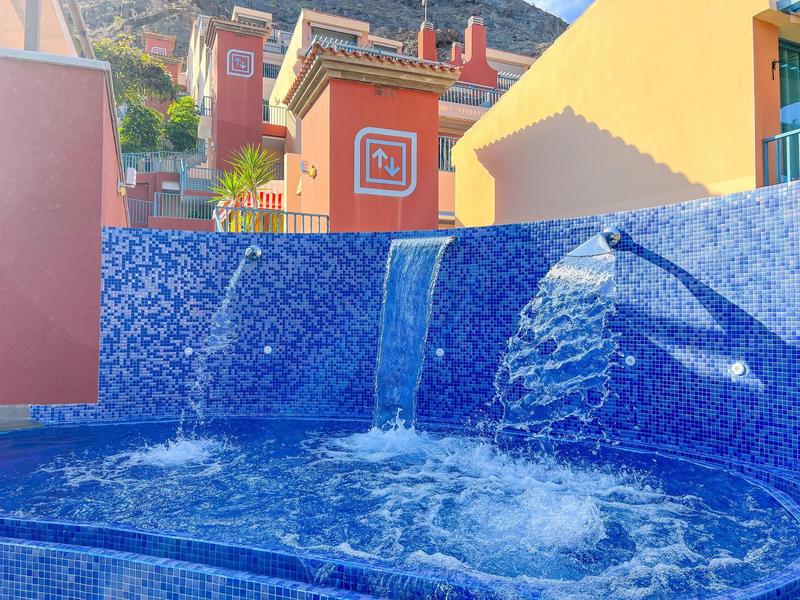 Blue tiled pool with waterfall and jet streams in a sunny courtyard with buildings.