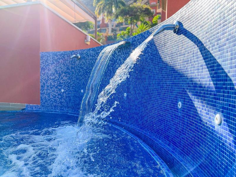 Blue tiled pool wall with water cascading from a curved spout under sunlight.