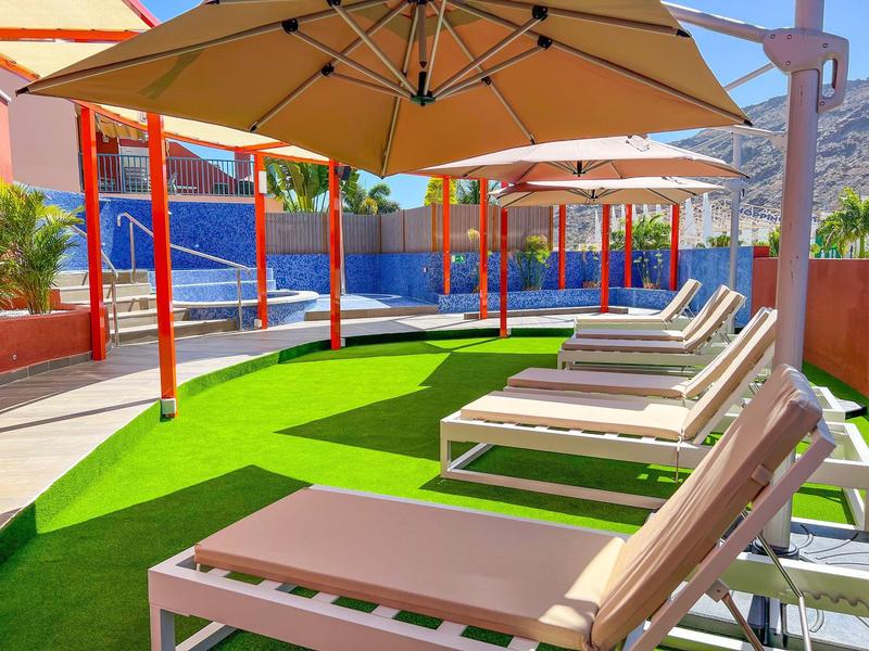Sun loungers under umbrellas by a pool on artificial grass in a sunny resort.