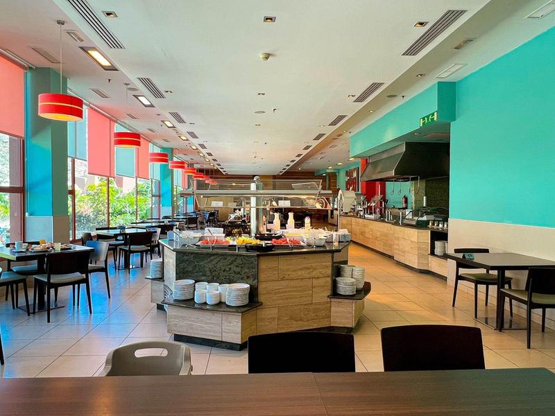 Bright hotel dining area with buffet counter, tables, chairs, and large windows with red blinds.