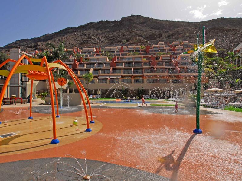 Splash park with orange climbing structure and water sprays by resort with hillside rooms