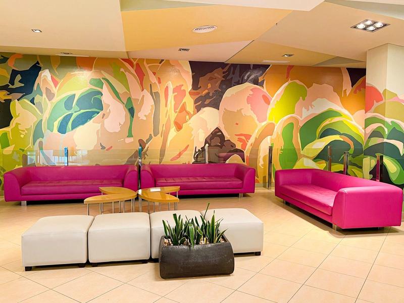 Bright lounge area with colorful floral mural, pink sofas, white seats, and plants on tiled floor.