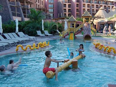 Kinder spielen im Pool mit Spielgeräten vor einem Hotel mit Liegestühlen und Grünpflanzen.