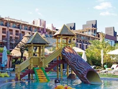 Bunter Kinderspielplatz mit Rutsche über Schwimmbecken in sonniger Hotelanlage.