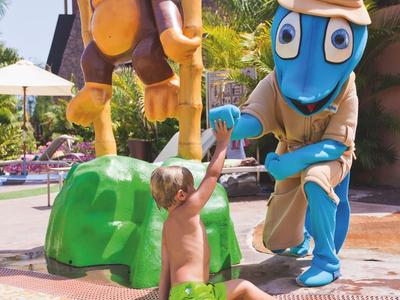 Kind spielt im flachen Pool neben großen bunten Figuren von Ente und Schlange mit Hut.