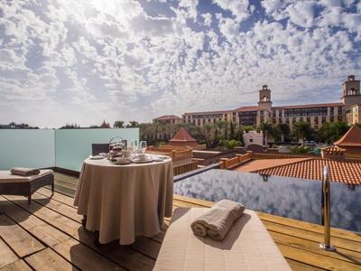Terrasse mit Holzboden, Tisch mit Decke, Stühlen und Liege vor Stadtkulisse und bewölktem Himmel.