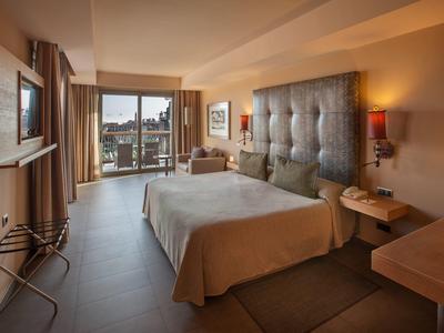 Helles Hotelzimmer mit großem Bett, Balkon, Fernseher, Sessel und warmen Beleuchtungselementen.