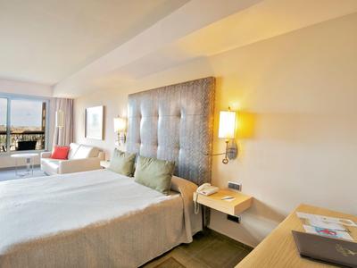 Helles Hotelzimmer mit großem Bett, gepolstertem Kopfteil, Nachtlampen und Fenster mit Ausblick.