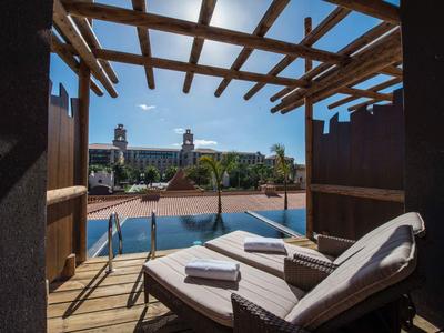 Blick von Holzbalkon mit Liege und Handtüchern auf Schwimmbad und historische Gebäude bei blauem Himmel.